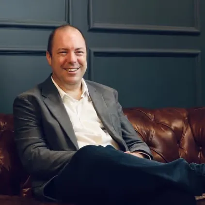 Smiling man in a gray blazer and white shirt sitting on a brown leather couch against a dark paneled wall.